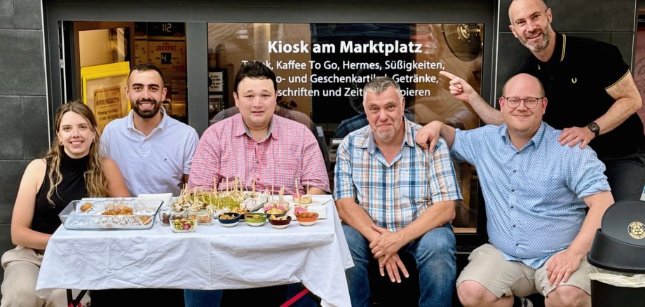 Fünf Männer und eine Frau vor einem Ladengeschäft sitzend. Davor ist ein Tisch mit Fingerfood platziert