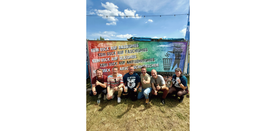 Fünf Männer und eine Frau knieend und lächelnd auf einem Acker vor einem bunten Banner.