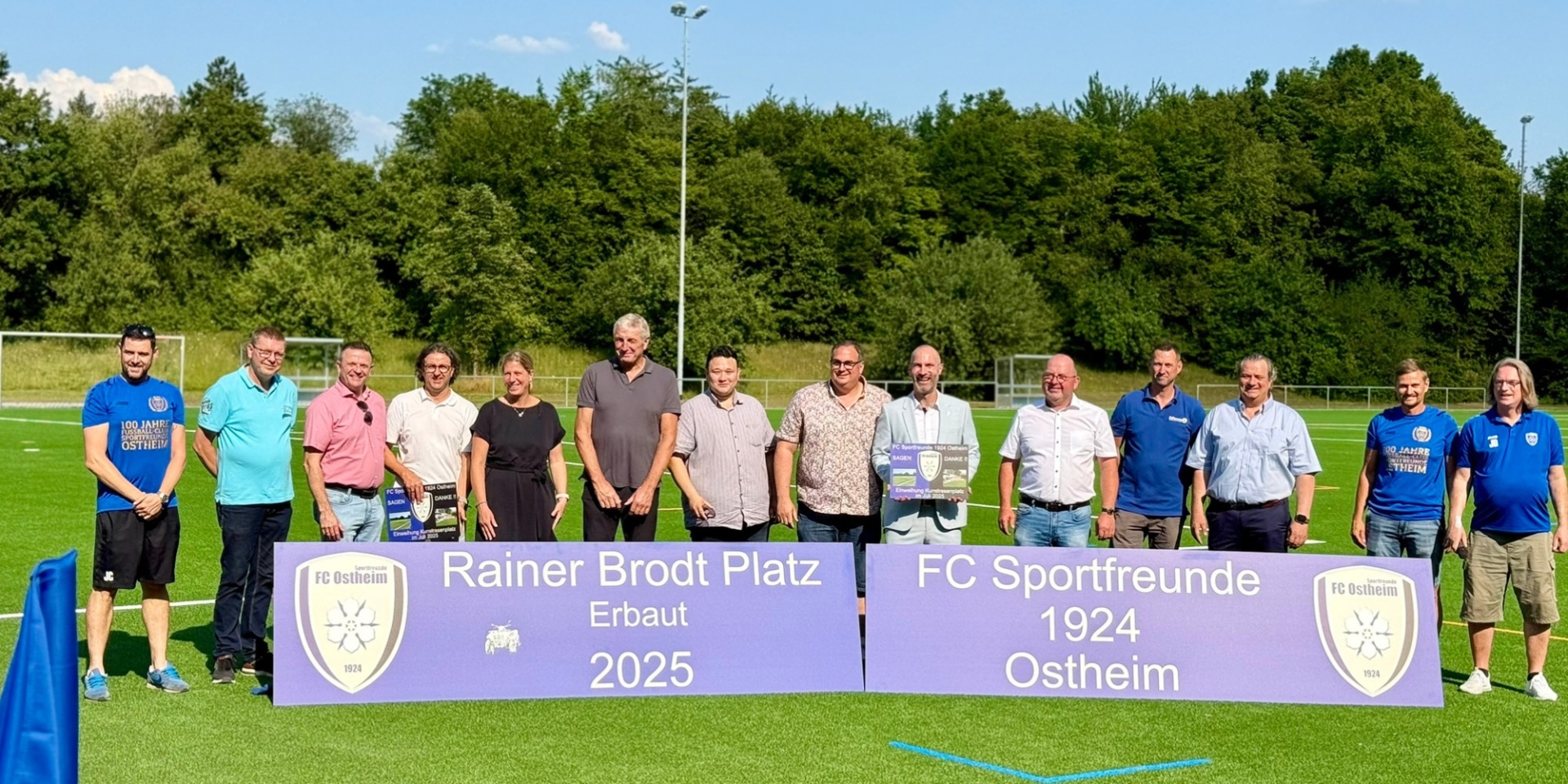 Mehrere Personen nebeneinander stehend auf einem Kunstrasenplatz/Sportfeld hinter einem Banner