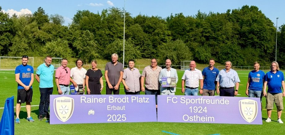 Mehrere Personen nebeneinander stehend auf einem Kunstrasenplatz/Sportfeld hinter einem Banner