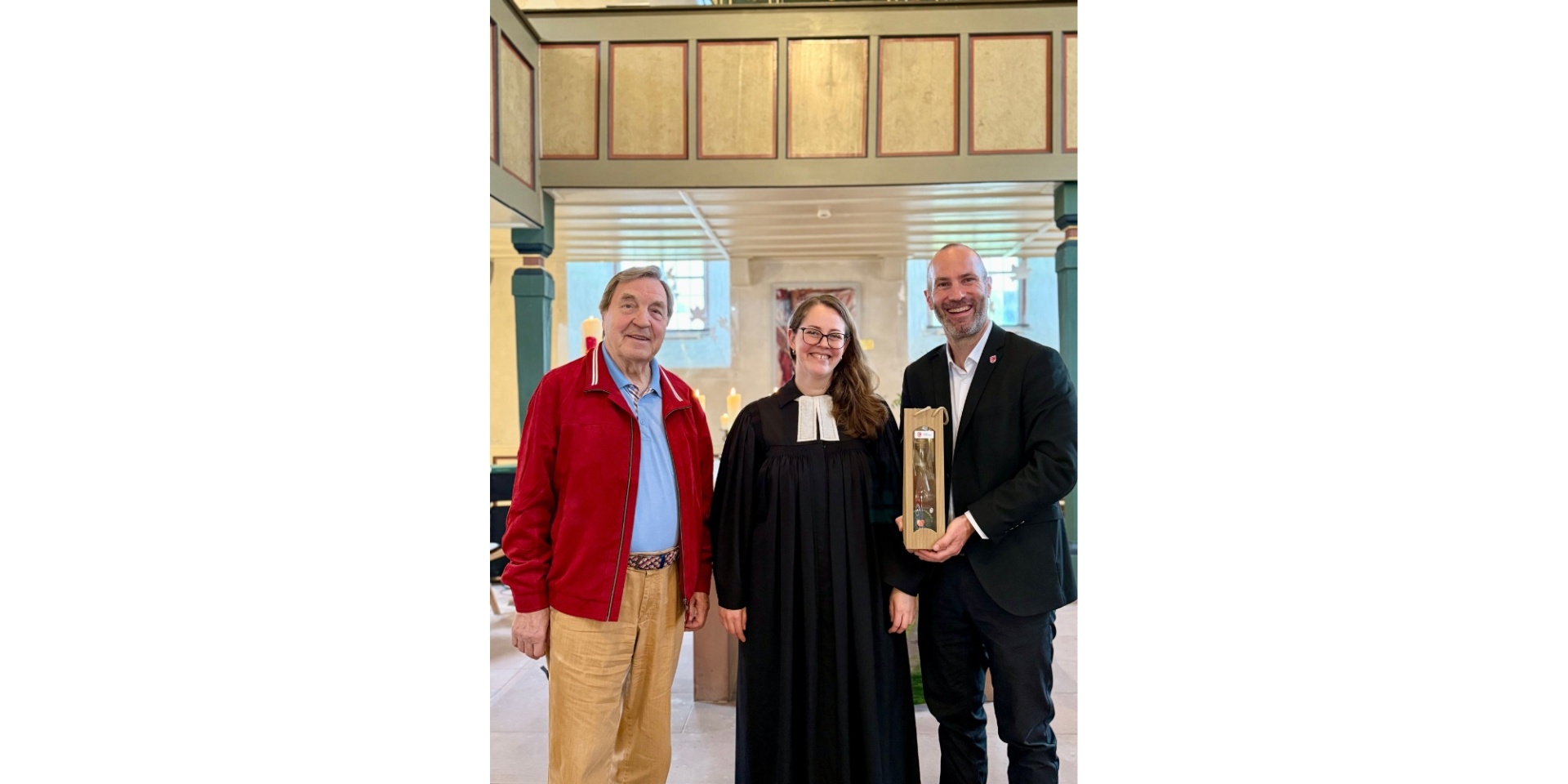 Eine Pfarrerin im schwarzen Talar zwischen zwei Männern stehend.