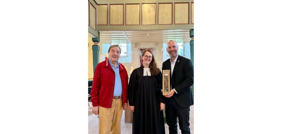 Eine Pfarrerin im schwarzen Talar zwischen zwei Männern stehend.