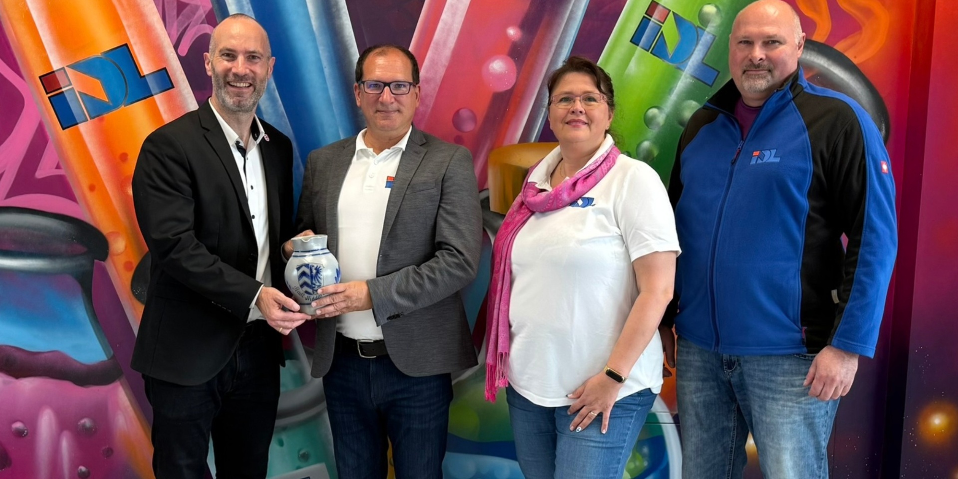 Drei Männer und eine Frau stehend vor einem bunten Hintergrund. Ein Mann überreicht einen Bembel