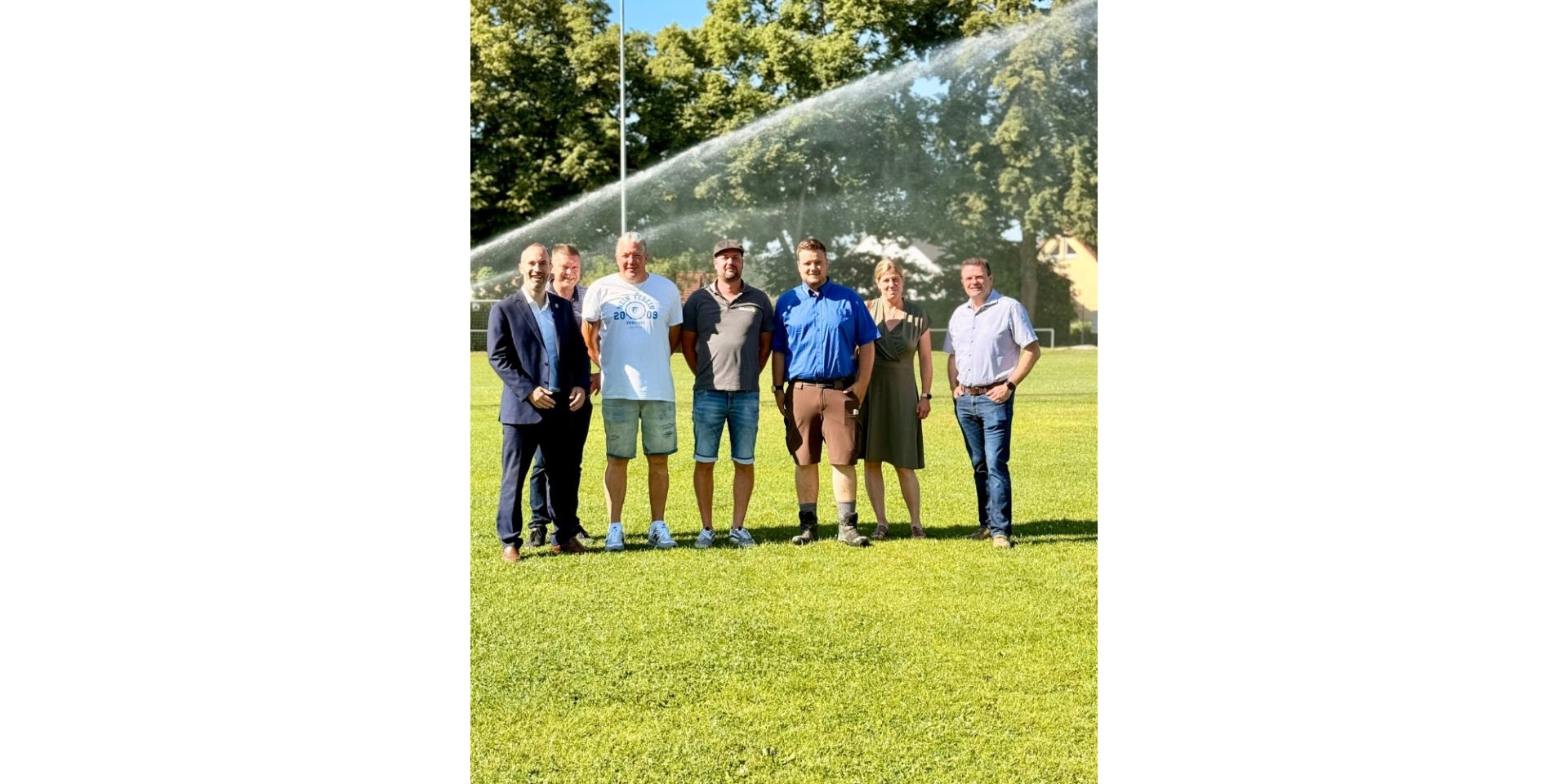 Mehrere Personen stehend auf einem Sportplatz. Im Hintergrund ist ein Wasserstrahl zu sehen