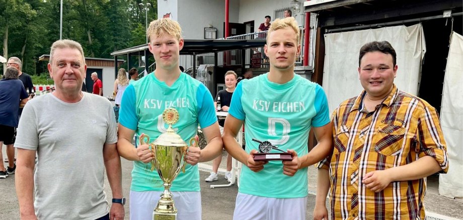 Vier Männer, zwei davon halten einen Pokal