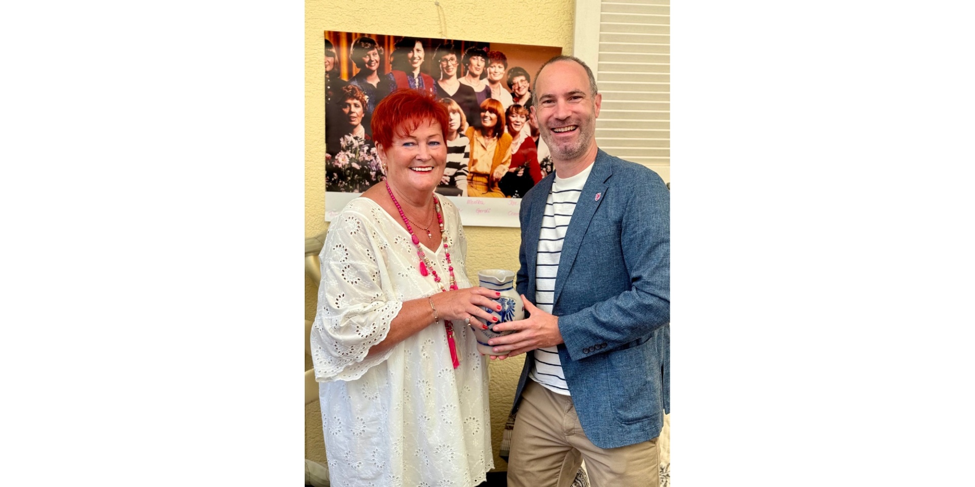 Eine Dame mit roten, kurzen Haaren nimmt einen Krug von einem Mann in einem blauen Jacket entgegen