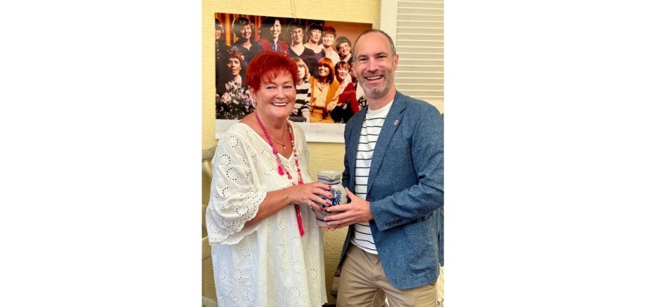 Eine Dame mit roten, kurzen Haaren nimmt einen Krug von einem Mann in einem blauen Jacket entgegen