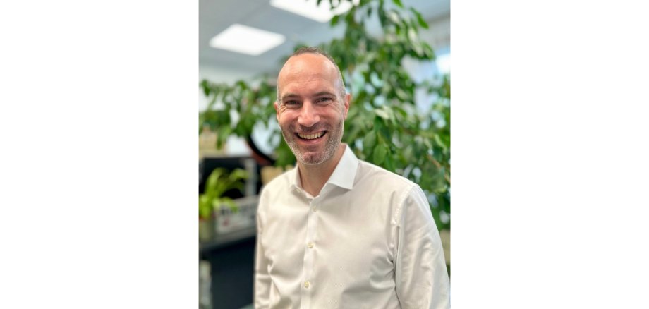 Mann in weißem Hemd lächelnd und stehend in einem Büro vor einer Grünpflanze