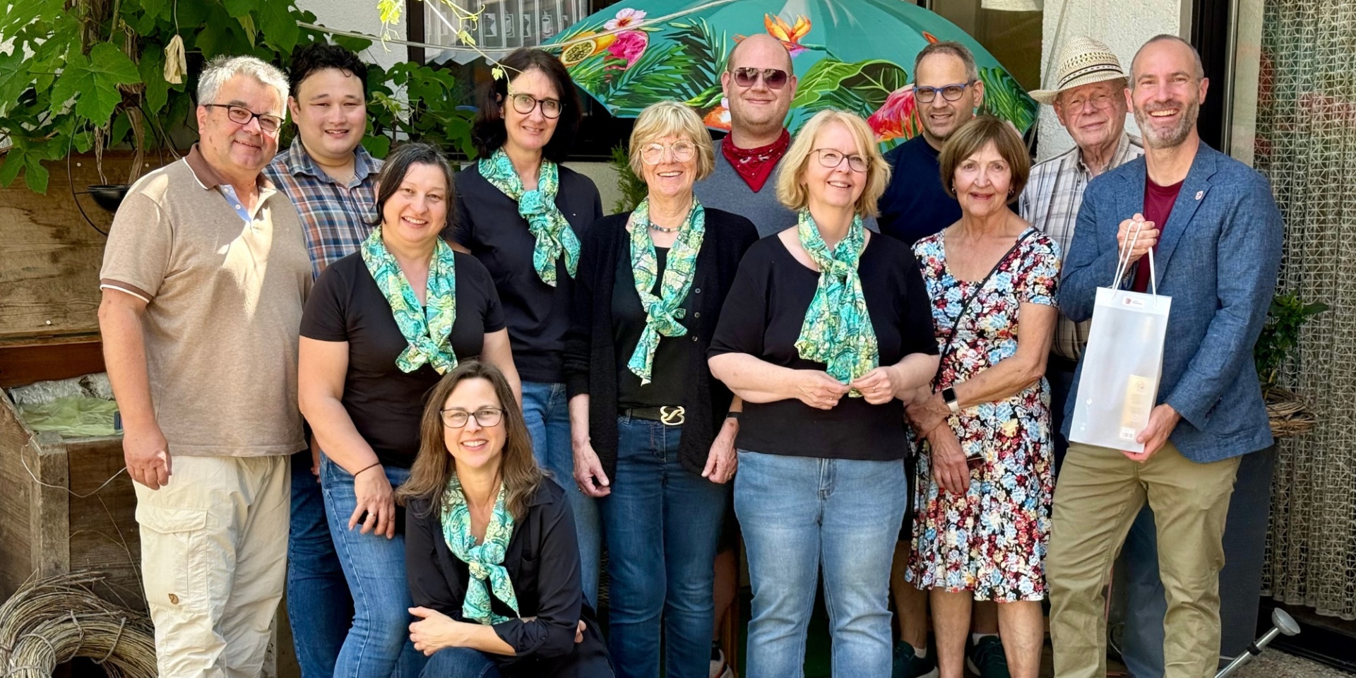 Mehrere Personen stehend vor einer Hausfassade. Davon tragen 4 Frauen schwarze Oberteile mit grün-blauen Halstüchern