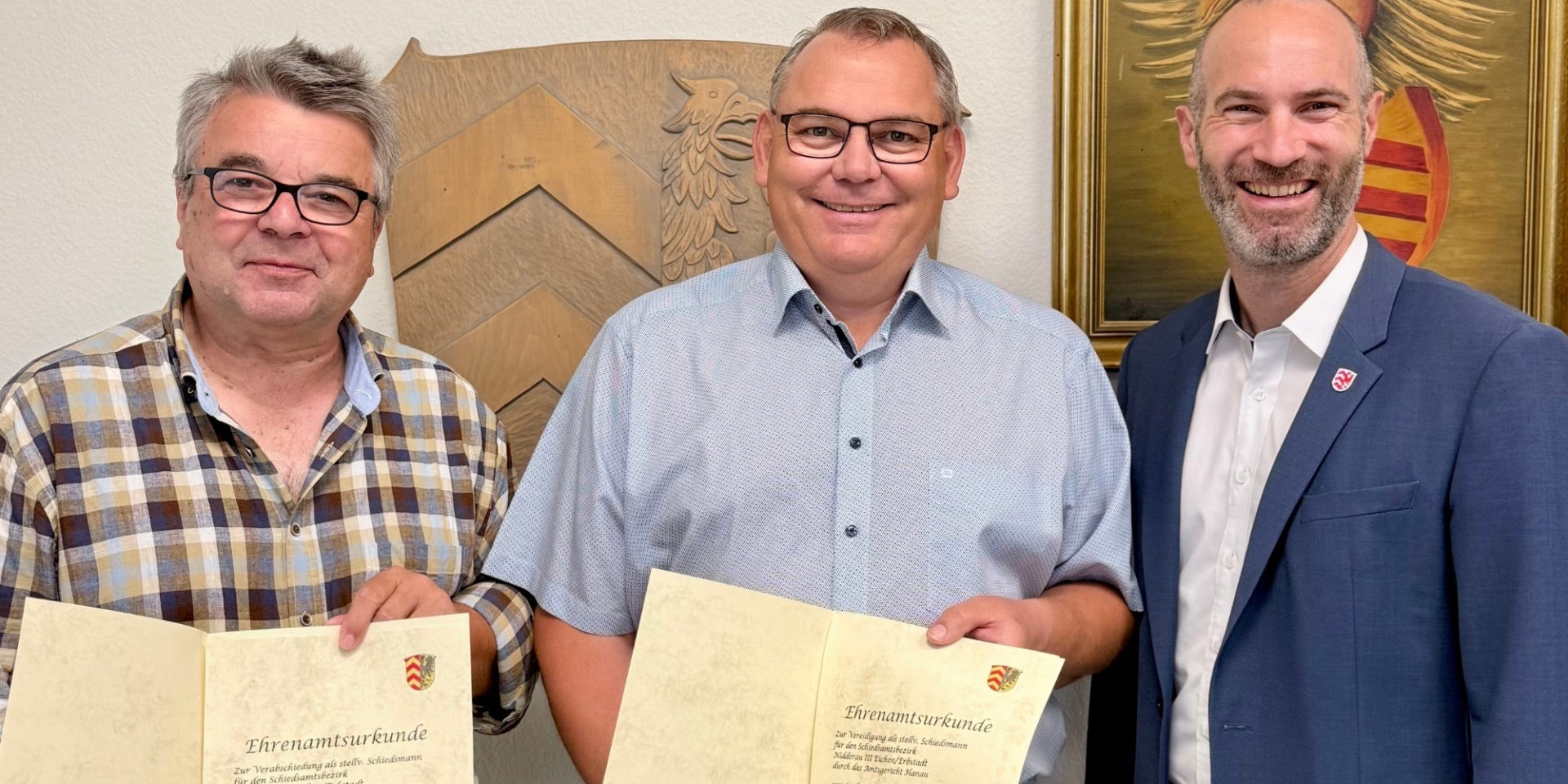 Drei Männer stehend nebeneinander. Zwei halten einen Ehrenamtsurkunde in den Händen