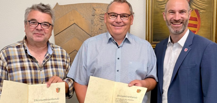 Drei Männer stehend nebeneinander. Zwei halten einen Ehrenamtsurkunde in den Händen