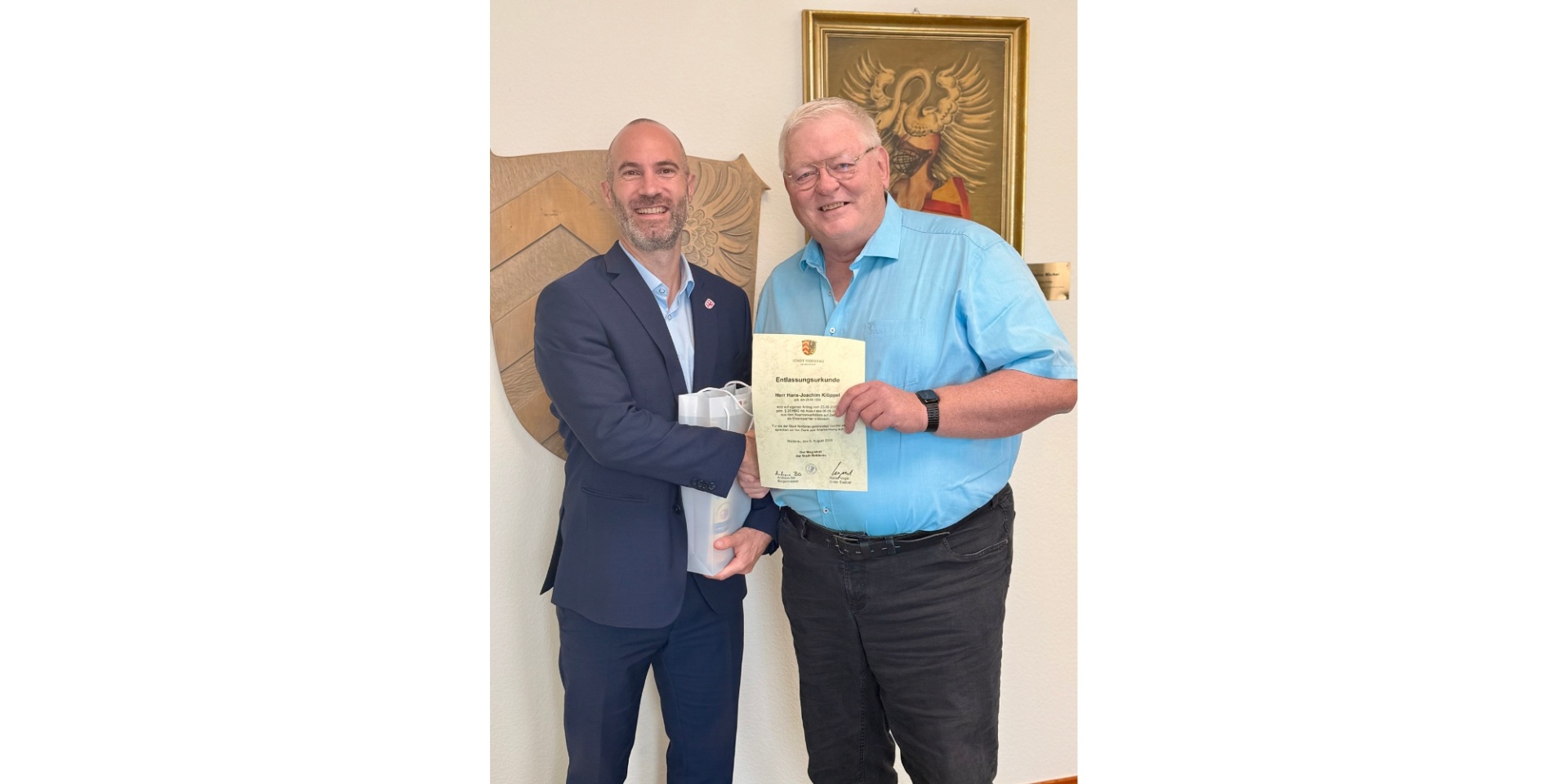 Zwei Männer stehend nebeneinander. Ein Mann hält eine Urkunde in der Hand