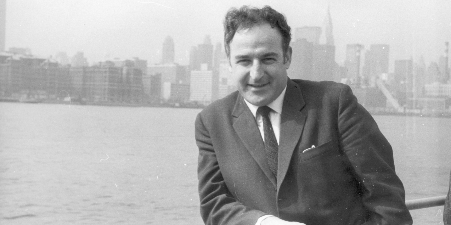 Schwarz-weiß-Aufnahme von einem Mann im Anzug lehnend an einem Geländer vor einem Fluss vor der Skyline von New York