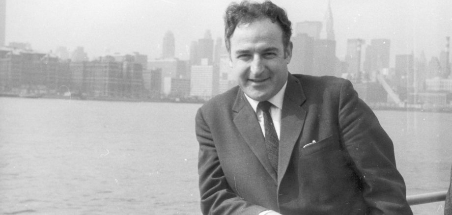 Schwarz-weiß-Aufnahme von einem Mann im Anzug lehnend an einem Geländer vor einem Fluss vor der Skyline von New York