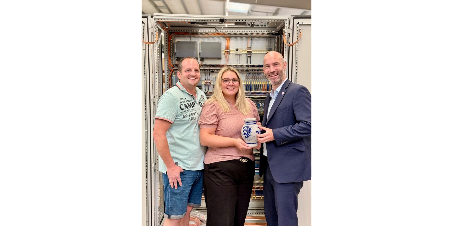 Zwei Männer und eine Frau stehend vor einer Schaltanlage. Die Frau hält einen hessischen Krug (Bembel) in den Händen