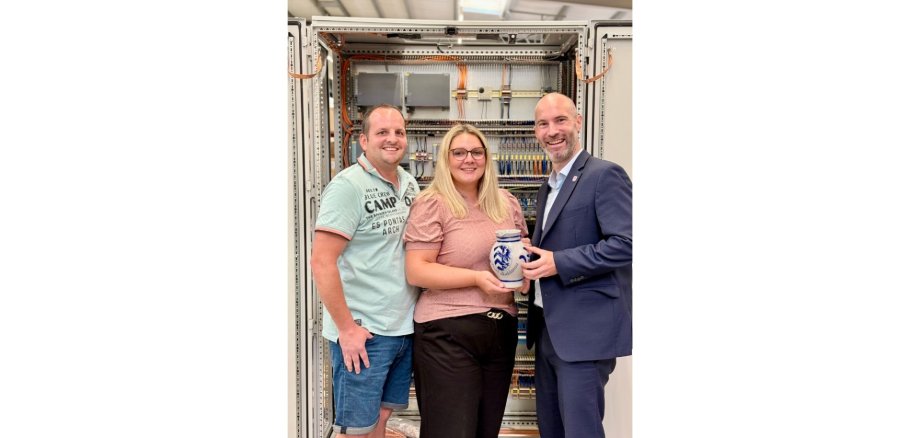 Zwei Männer und eine Frau stehend vor einer Schaltanlage. Die Frau hält einen hessischen Krug (Bembel) in den Händen