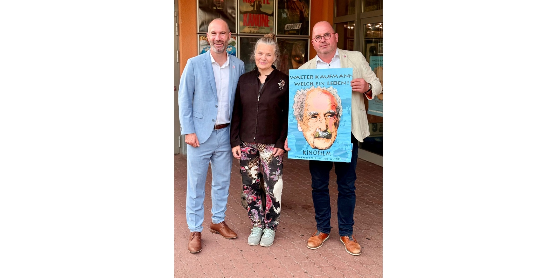 Zwei Männer und eine Frau stehend vor einem Gebäude. Ein Mann hält ein Poster in der Hand