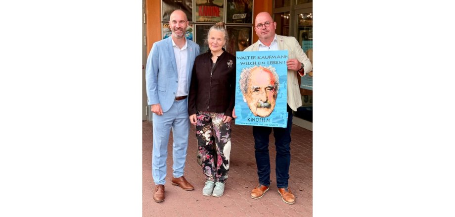 Zwei Männer und eine Frau stehend vor einem Gebäude. Ein Mann hält ein Poster in der Hand