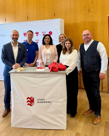 Fünf Personen stehend hinter einem weißen Rednerpult mit rotem Wappen