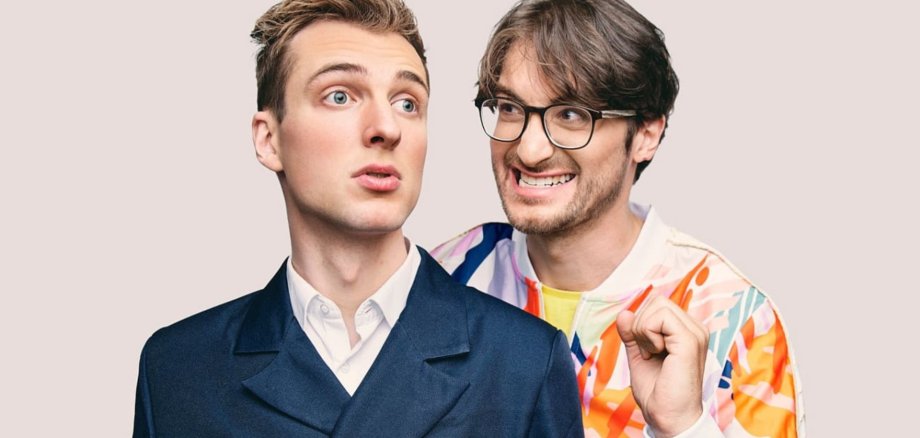 Zwei Männer nebeneinander. Einer trägt einen dunkelblauen Anzug, der andere einen bunten Anzug