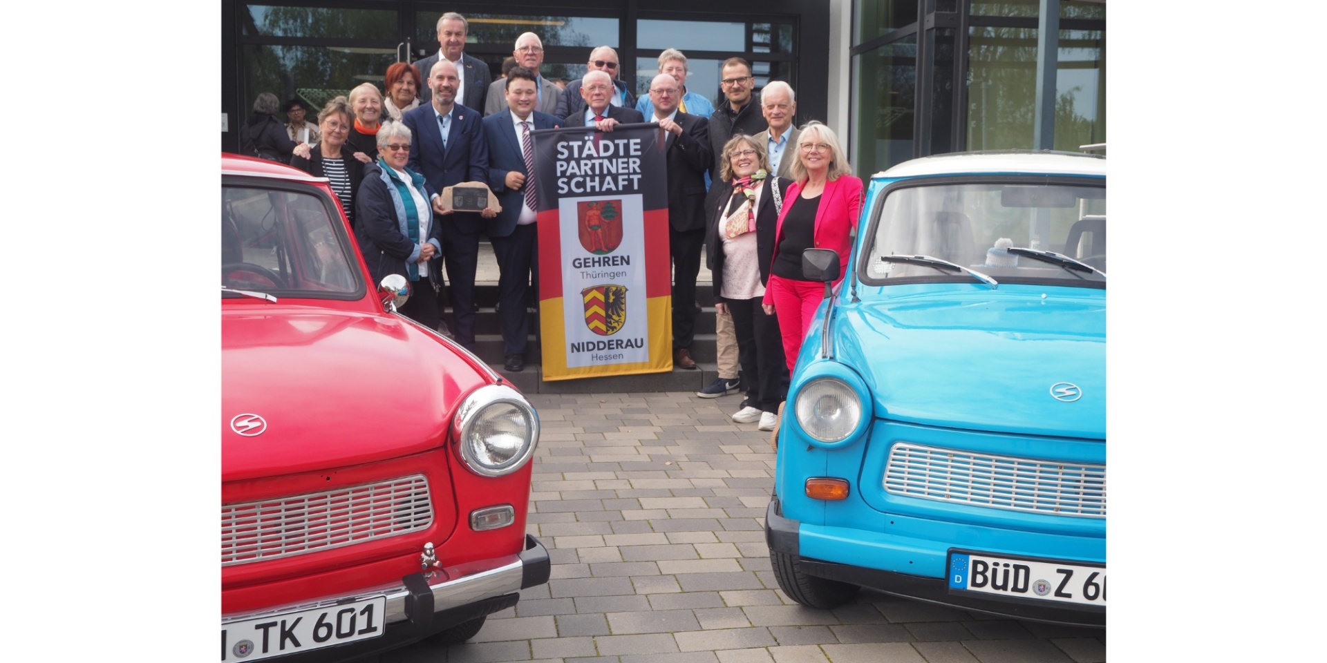 Eine Personengruppe zusammenstehend mittig. Links und rechts jeweils ein Auto.