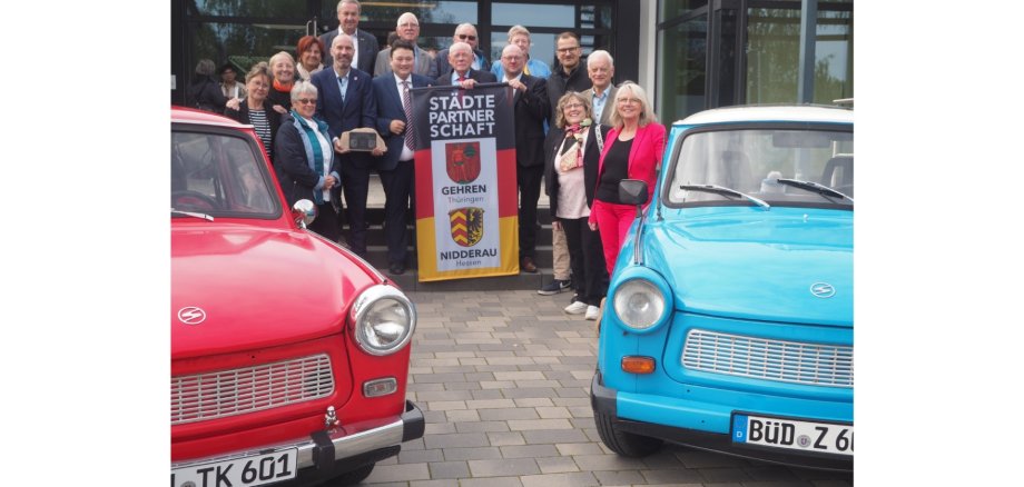 Eine Personengruppe zusammenstehend mittig. Links und rechts jeweils ein Auto.