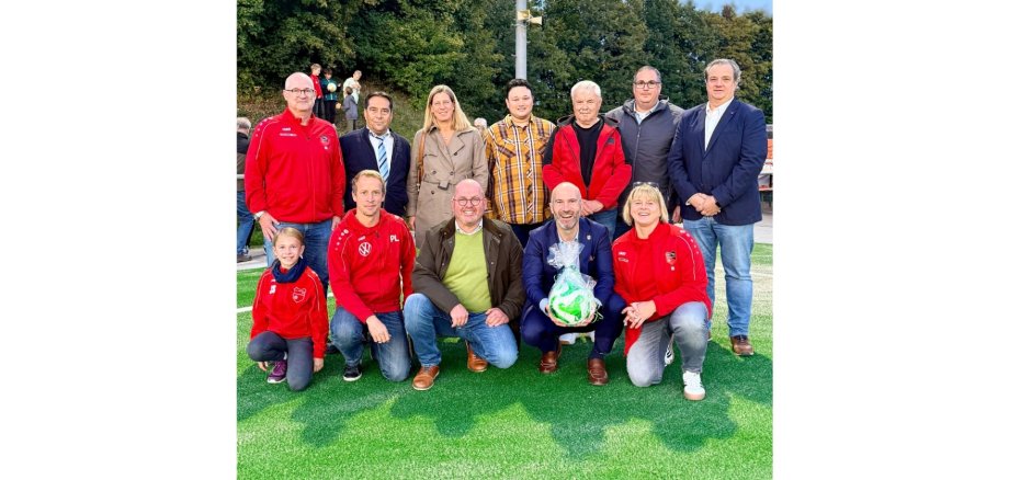 Eine Personengruppe zusammenstehend auf einem Kunstrasen. Ein Mann hält einen verpackten Fußball in der Hand.