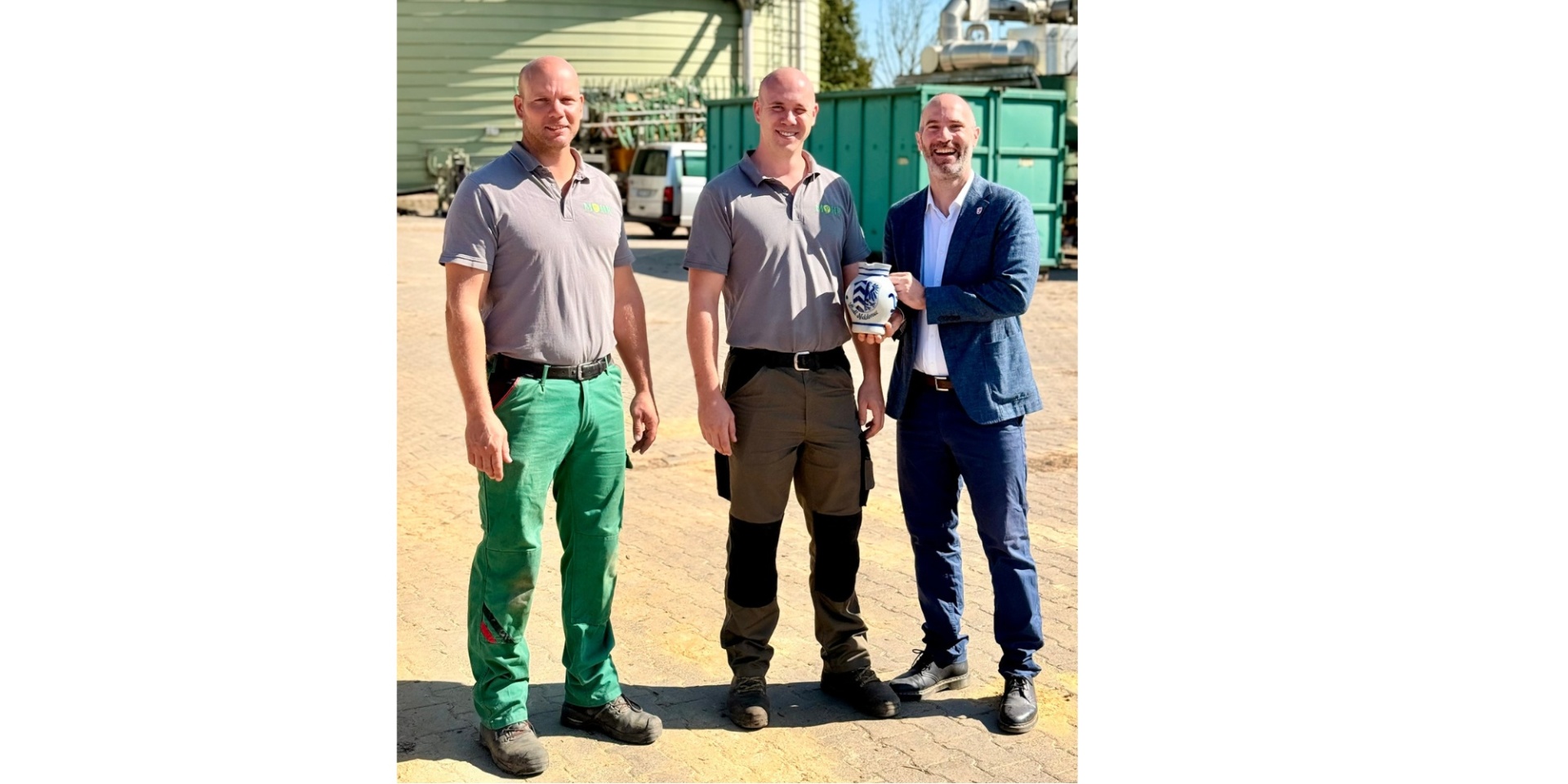 Drei Männer stehend vor einem Industriehintergrund. Ein Mann hält einen Bembel in der Hand.