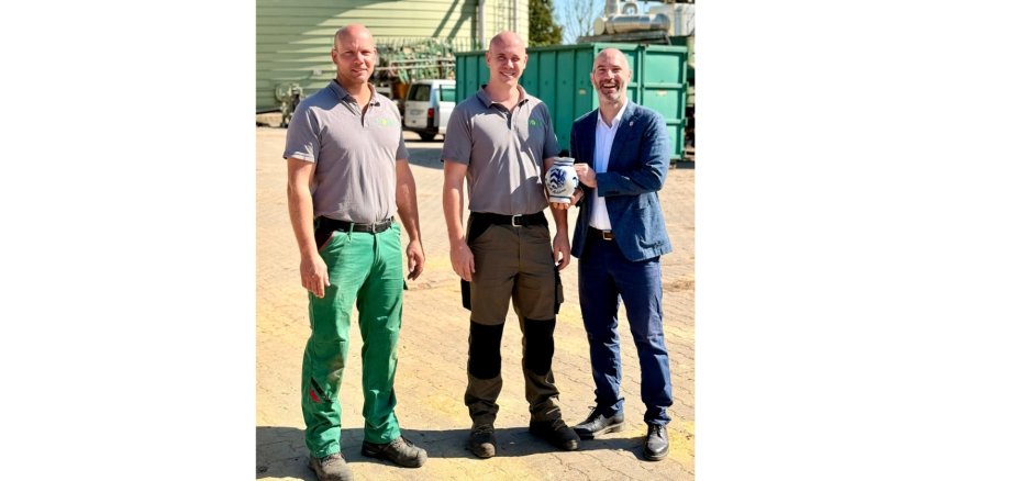 Drei Männer stehend vor einem Industriehintergrund. Ein Mann hält einen Bembel in der Hand.