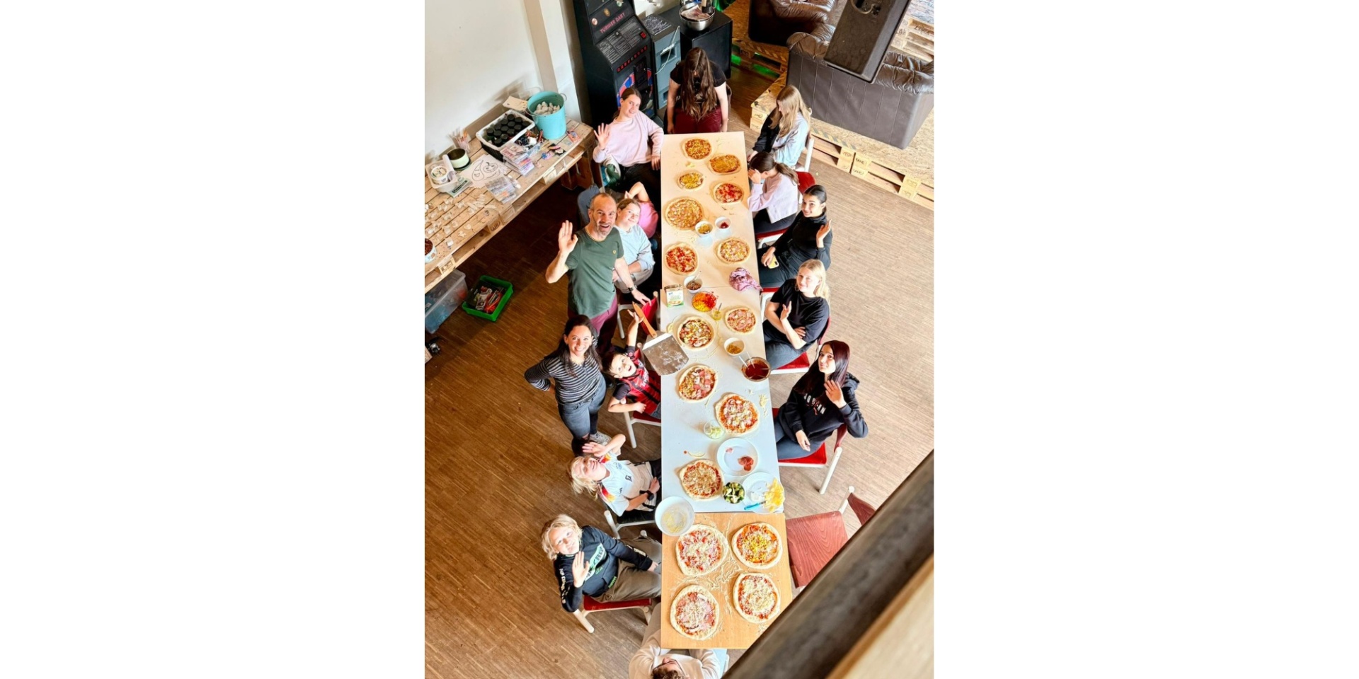 Mehrere Kinder und 1 Mann bereiten Pizzen an einem langen großen Tisch vor