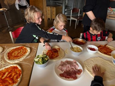 3 Kinder sitzen an einem Tisch und bereiten Pizzen vor mit verschiedenen Belägen