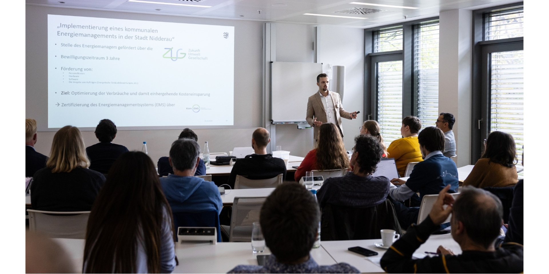 Ein Mann stehend vor einer sitzenden Personengruppe an Tischen. Im Hintergrund eine Abbildung einer Präsentation.