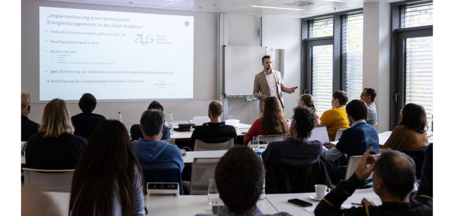 Ein Mann stehend vor einer sitzenden Personengruppe an Tischen. Im Hintergrund eine Abbildung einer Präsentation.