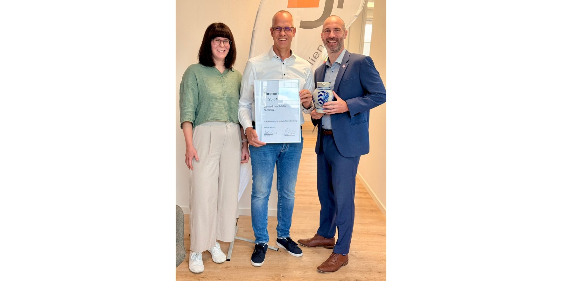 Zwei Männer und eine Frau stehend. Der eine Mann im dunklen Anzug hält einen Krug in der Hand