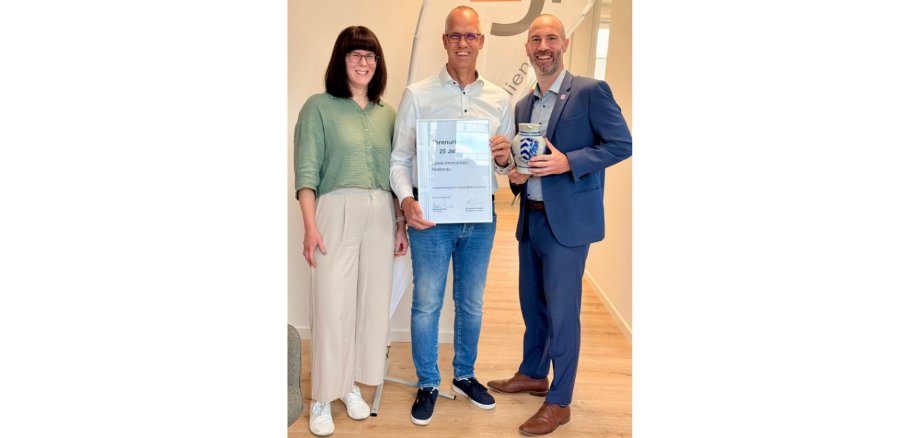 Zwei Männer und eine Frau stehend. Der eine Mann im dunklen Anzug hält einen Krug in der Hand