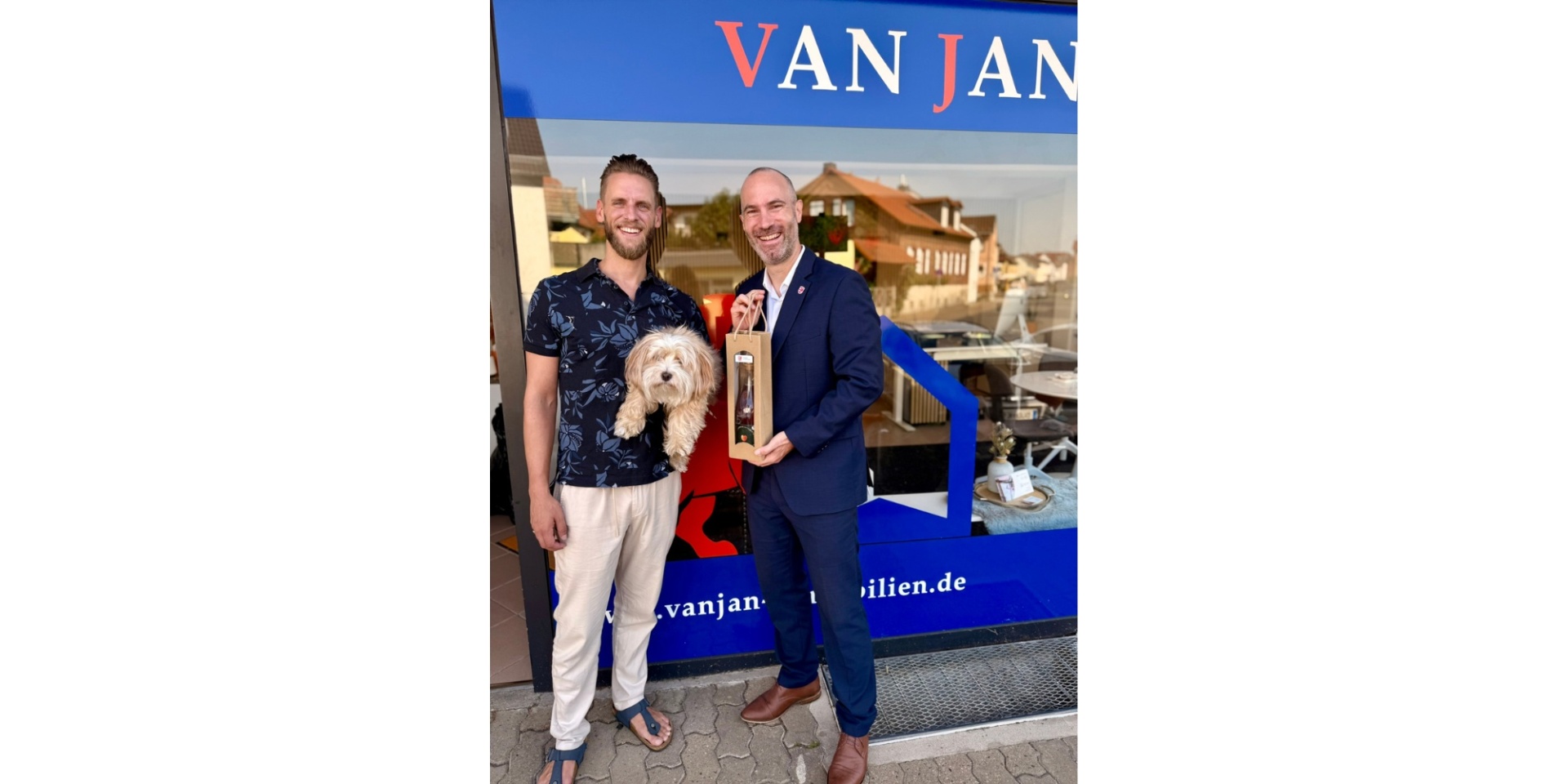 Zwei Männer stehend vor einem Geschäft. Der eine Mann hält einen kleinen Hund auf dem Arm