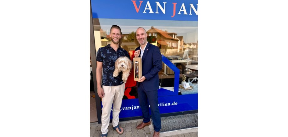 Zwei Männer stehend vor einem Geschäft. Der eine Mann hält einen kleinen Hund auf dem Arm