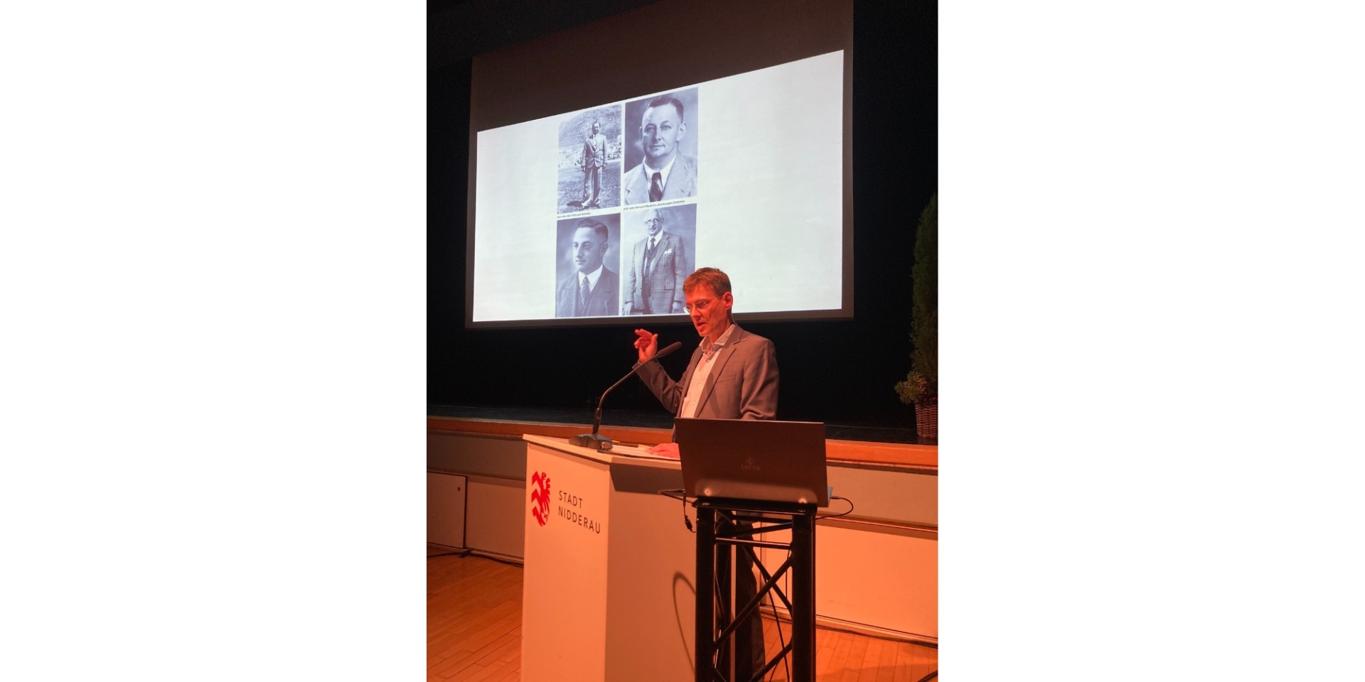 Mann stehend hinter einem Rednerpult auf einer Bühne. Hinter ihm ist eine Leinwand aufgebaut mit schwarz-weiß Fotos