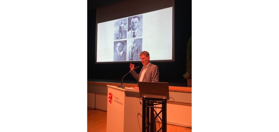 Mann stehend hinter einem Rednerpult auf einer Bühne. Hinter ihm ist eine Leinwand aufgebaut mit schwarz-weiß Fotos