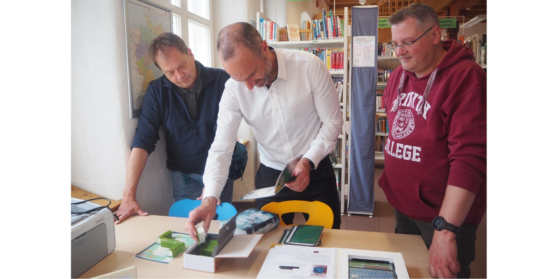 Drei Männer stehend und konzentriert auf eine Schachtel auf einem Tisch schauend.