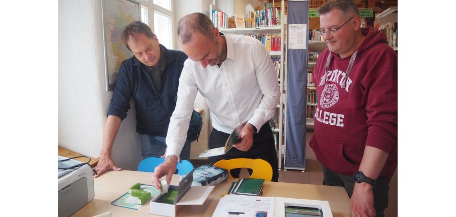 Drei Männer stehend und konzentriert auf eine Schachtel auf einem Tisch schauend.