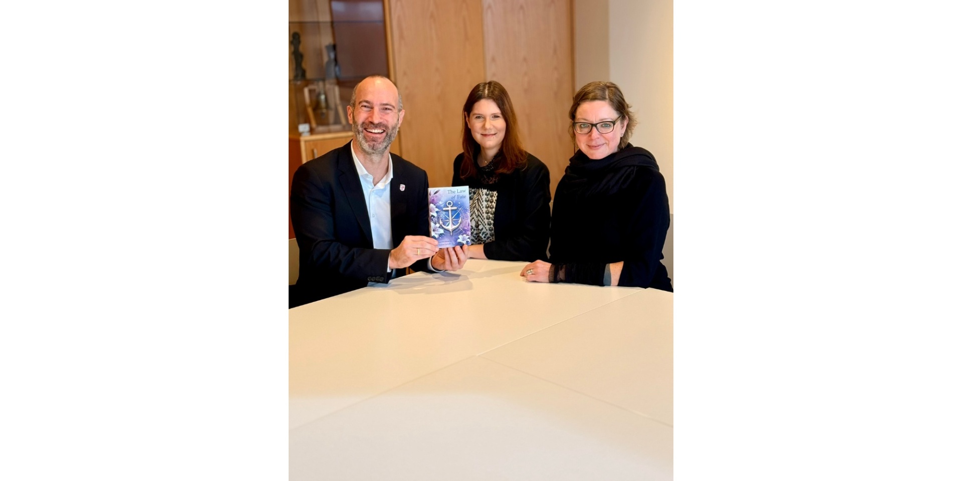 Ein Mann mit zwei Frauen an einem Tisch sitzend. Der Mann hält ein Buch in der Hand