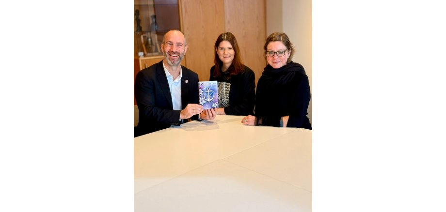 Ein Mann mit zwei Frauen an einem Tisch sitzend. Der Mann hält ein Buch in der Hand