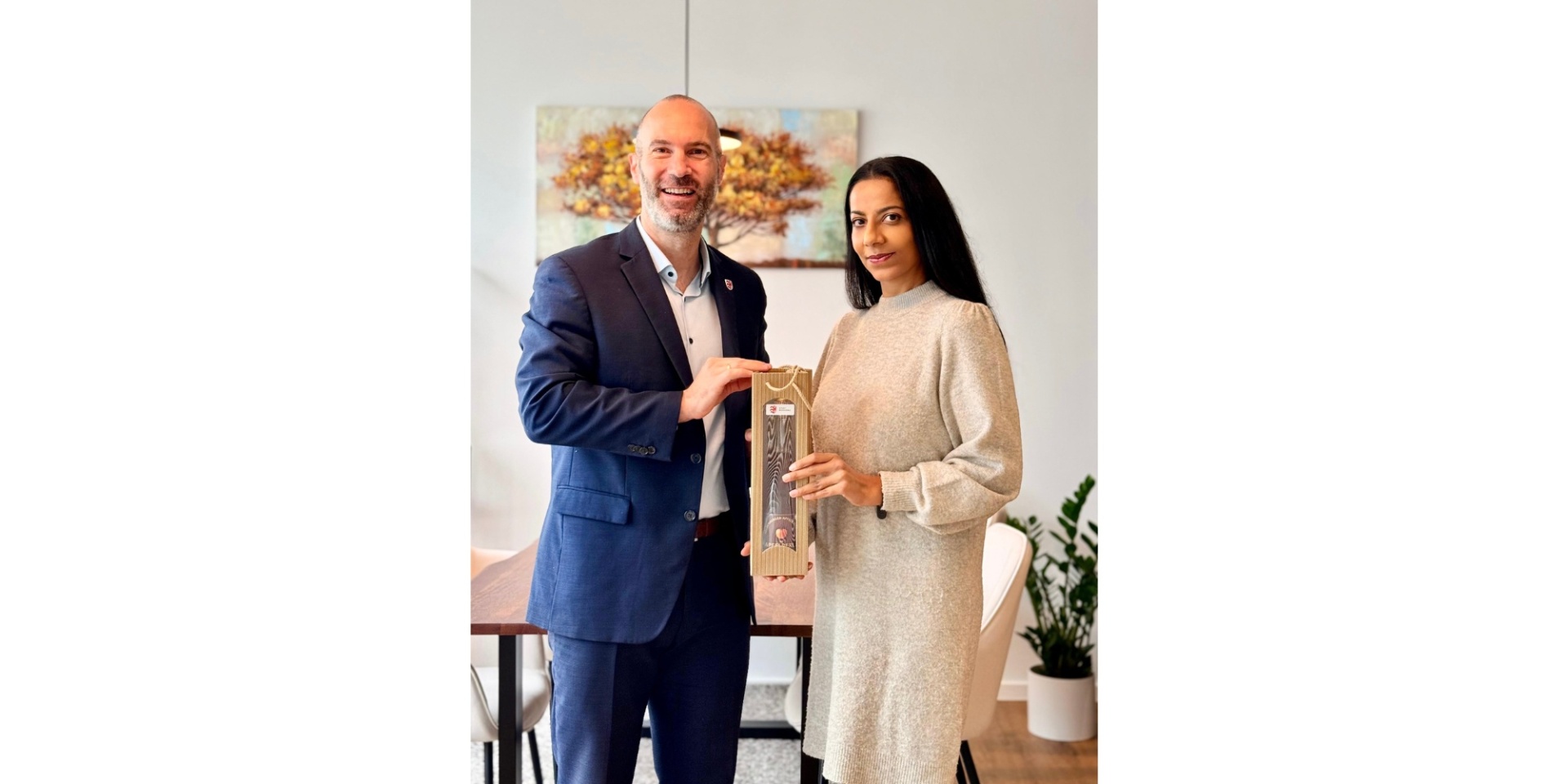 Ein Mann und eine Frau stehend. Die Frau erhält eine Flasche als Geschenk überreicht.