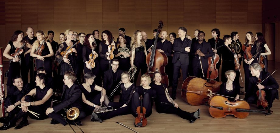 Mehrere Personen eines Orchesters mit Musikinstrumenten haltend vor einem schwarzen Hintergrund