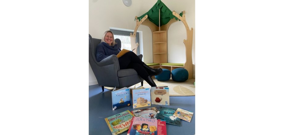 Frau sitzend in einem gemütlichen Stuhl in einer Kita. Davor sind mehrere Kinderbücher liegend auf dem Boden