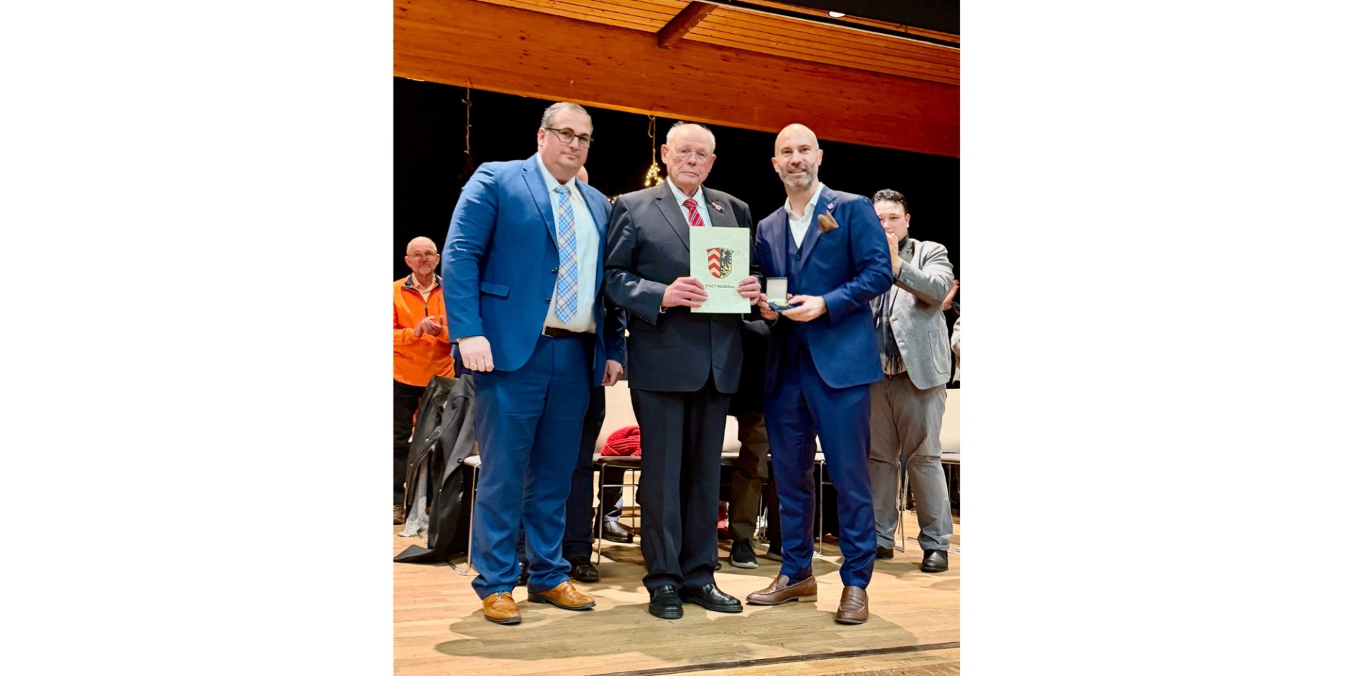 Drei Männer stehend zusammen auf einer Bühne. Der Mann in der Mitte hält eine Urkunde in den Händen