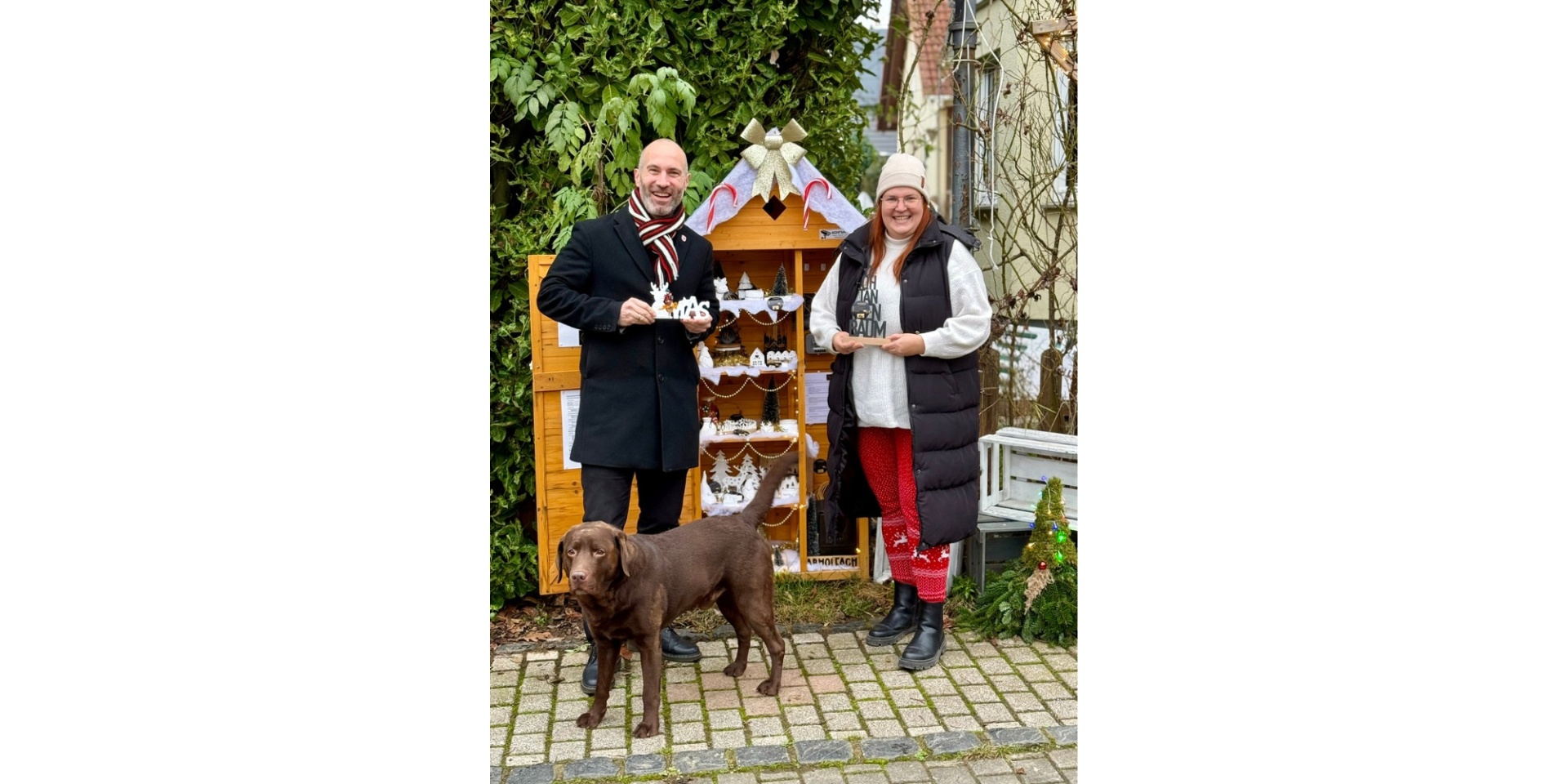 Ein Mann und eine Frau stehend vor einem Schrank mit gebastelten Werken. Davor steht ein dunkler Hund