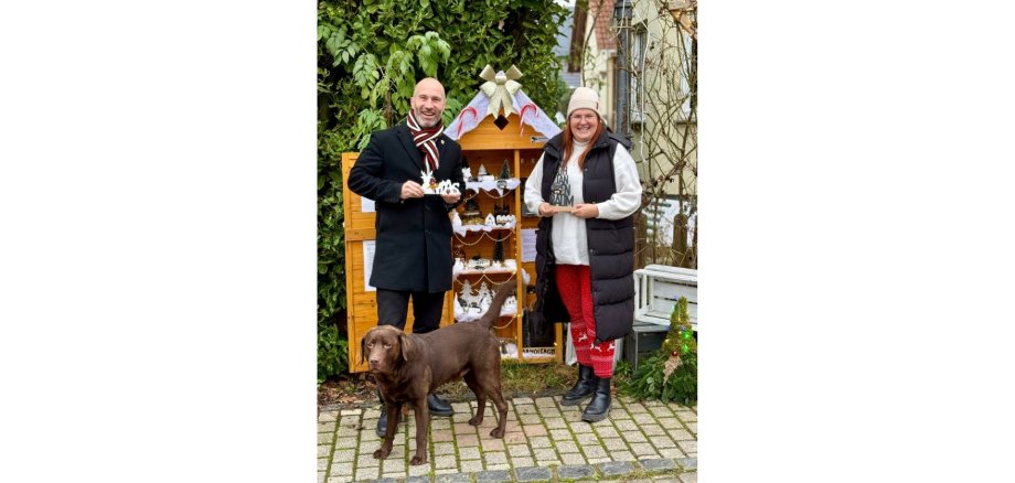 Ein Mann und eine Frau stehend vor einem Schrank mit gebastelten Werken. Davor steht ein dunkler Hund