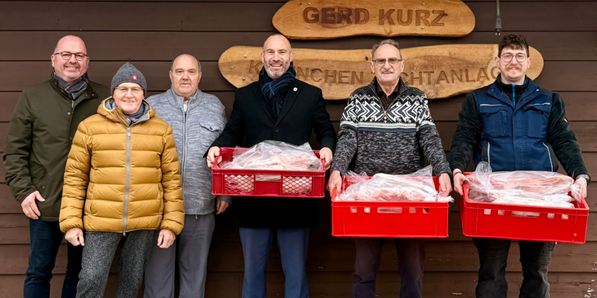 Sechs Männer stehend nebeneinander vor einem Holzhaus. Drei Männer halten roten Kisten in den Händen